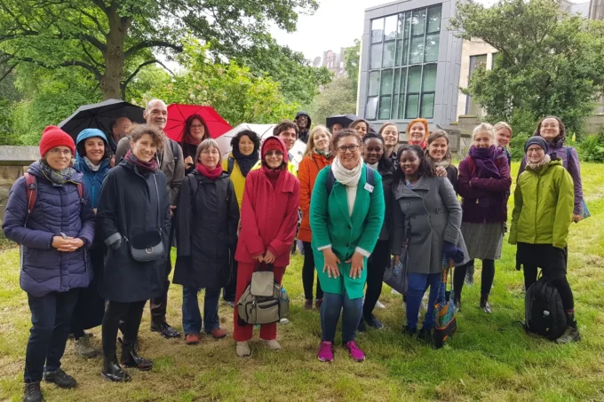 Group of people in autmn and winter coats, some with umbrellas, smiling at the camera. They're standing on a grassy lawn with trees and buildings in the background.