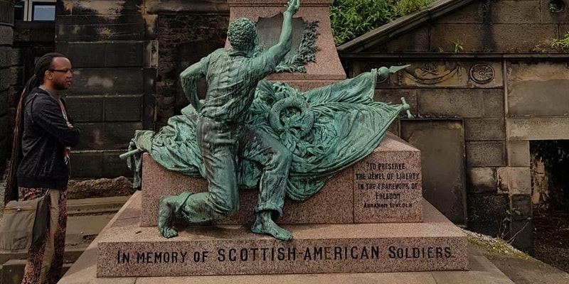 Black man stands looking at a statue. Statue is a figure leaning on flags. Stone is engraved with text: 'In memory of Scottish-American soldiers'