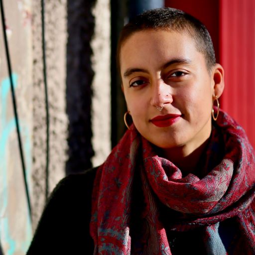 Brown woman with shaved head looks at the camera. There is bright sunlight and she is wearing a scarf.