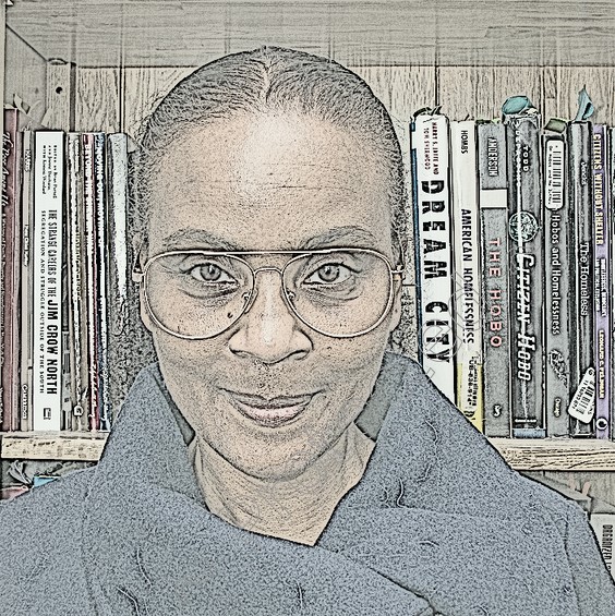 Black woman standing in front of a bookcase is smiling. She wears a wrap-neck coat and glasses and her hair is tied back.