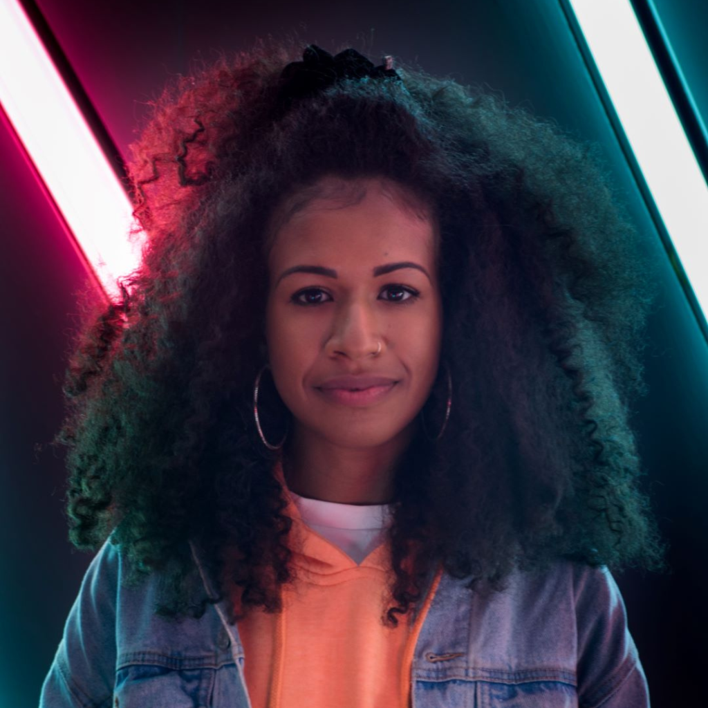 Person with long afro hair, wearing an orange and blue top and smiling at the camera in front of neon lights.