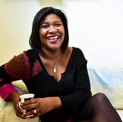 Photo of a Black woman, sitting on a sofa, smiling, laughing warmly, holding a mug. She is wearing a dark knitted top.