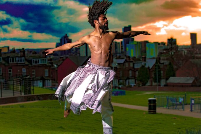 Photo of a Black person dancing, arms outstretched and locks flowing; wearing a white and patterned costume.