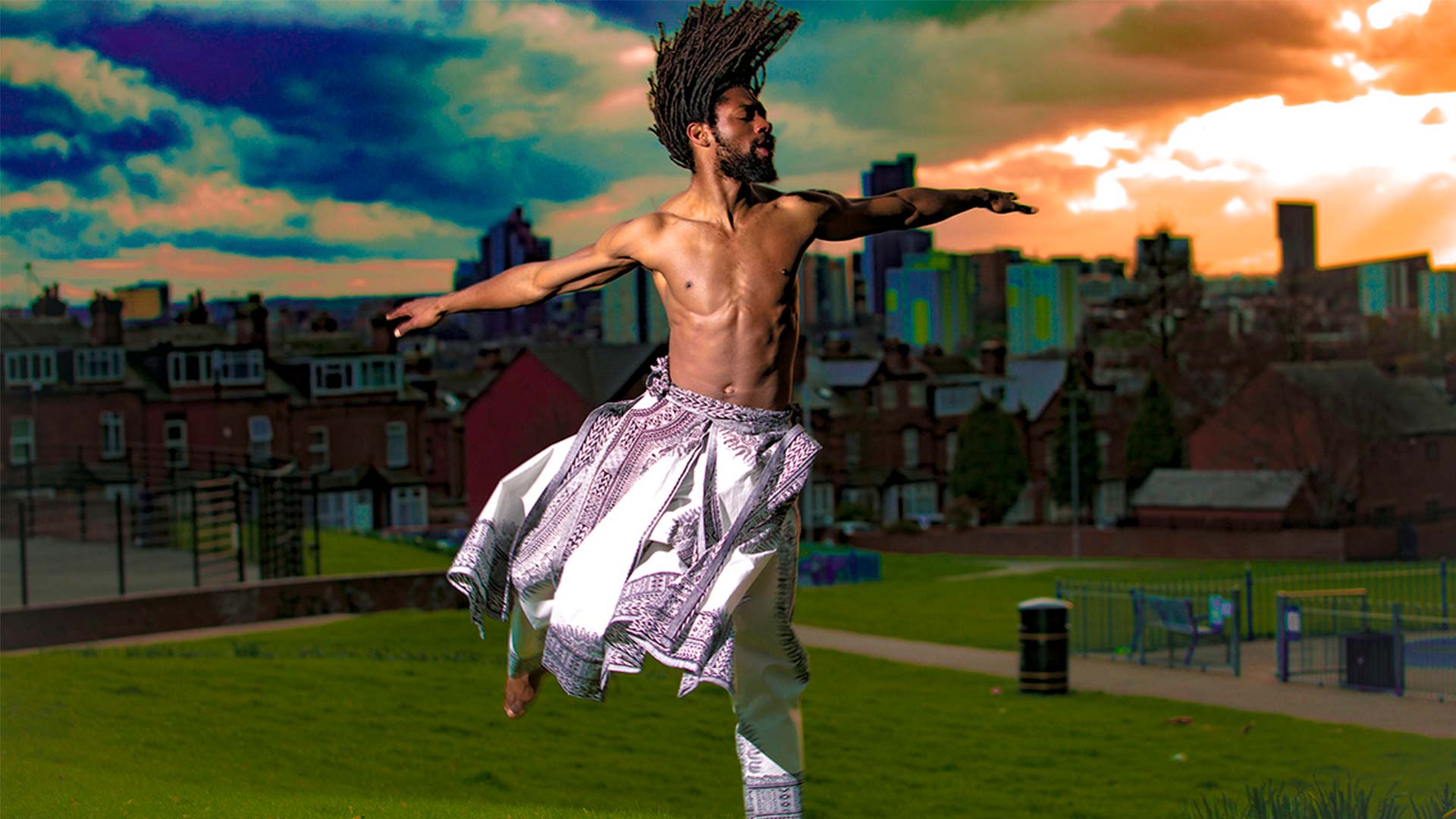 Photo of a Black person dancing, arms outstretched and locks flowing; wearing a white and patterned costume.