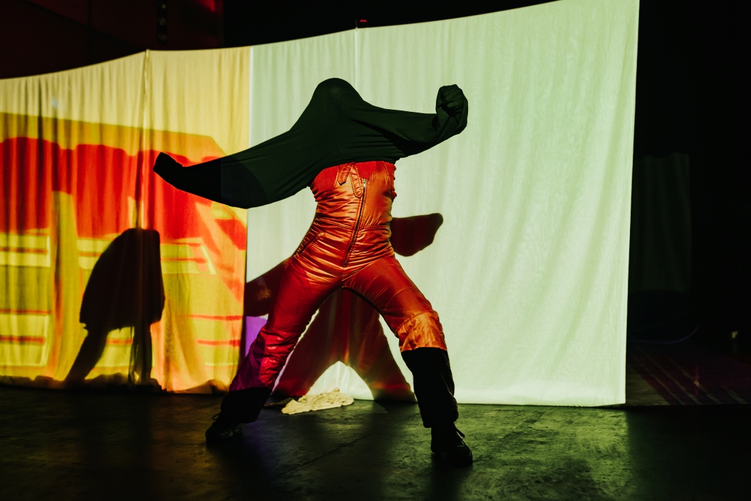 Black person wearing a red jumpsuit has a piece of black fabric on their head, blocking their vision and connecting to their hands, creating the feeling of tension.