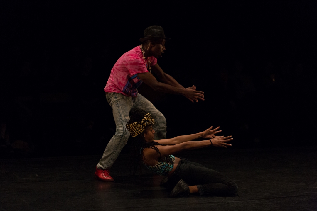 There is two Black dancers - one of them is standing and the other one is on their knees. They both have their hands stretched, reaching in front of them.