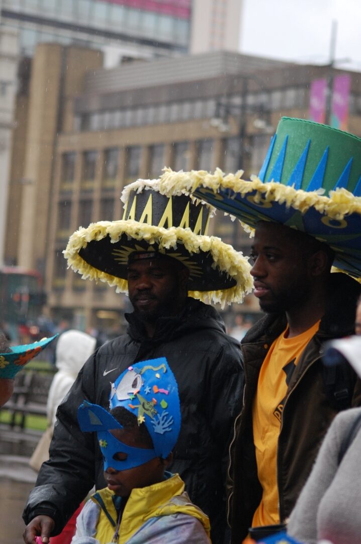 Two Black people in a Carnival setting are wearing big colourful hats. There's also a young black person in front of them wearing a blue Carnival mask.