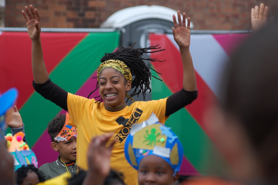 A Black adult person is jumping amongst black children while smiling. The adult is wearing a yellow t-shirt and the children are wearing carnival masks and costumes. 