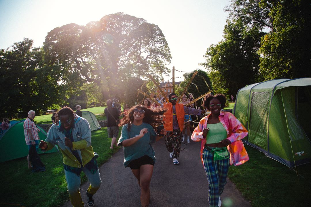 Four Black people are dancing joyously along a park path. One person is wearing a carnival mas costume which radiates outward in golden rays. There are tents either side of the path and some onlookers.