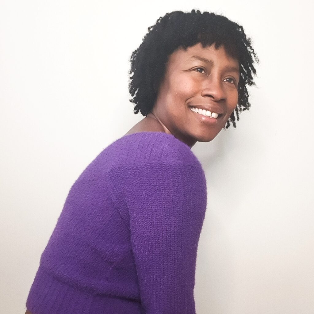 Person wearing a purple jumper, turned sideways and looking at the camera and smiling.