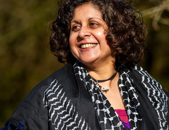 Photo of Anthony V. Capildeo, smiling and looking towards the left, wearing a black and white keffiyeh over an orange top.