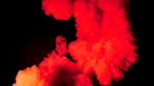 A photo of a dancer in warm red light lighting, looking serenely to the left, wearing a costume of cloud-like ruffles.