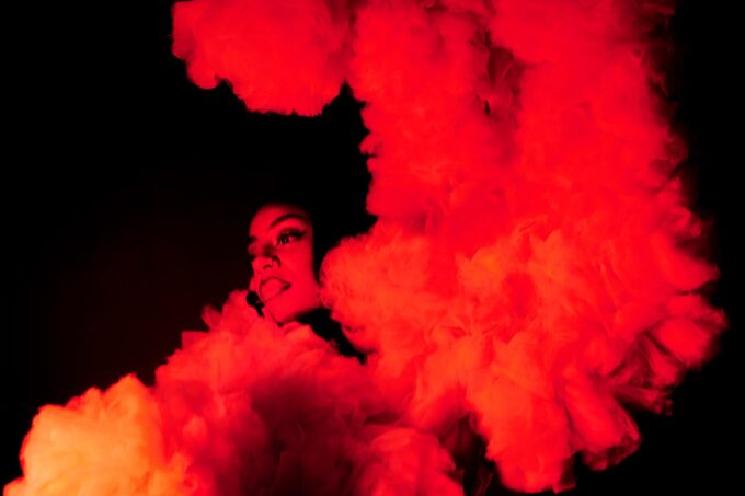 A photo of a dancer in warm red light lighting, looking serenely to the left, wearing a costume of cloud-like ruffles.