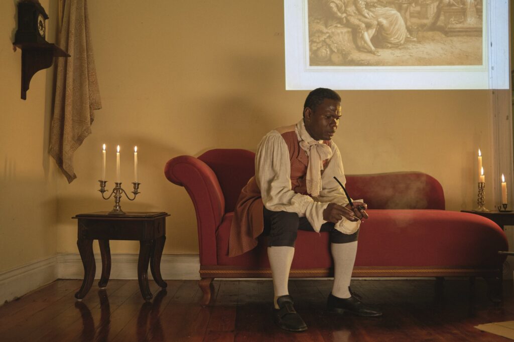 Film still. A Black man in 18th Century clothes sits on a chaise lounge, holding a pipe, thinking deeply. There is a side table with a candelabra to the left.