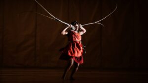 Photo of dancer performing, dressed in a red smock and holding long antler-like props by their ears.