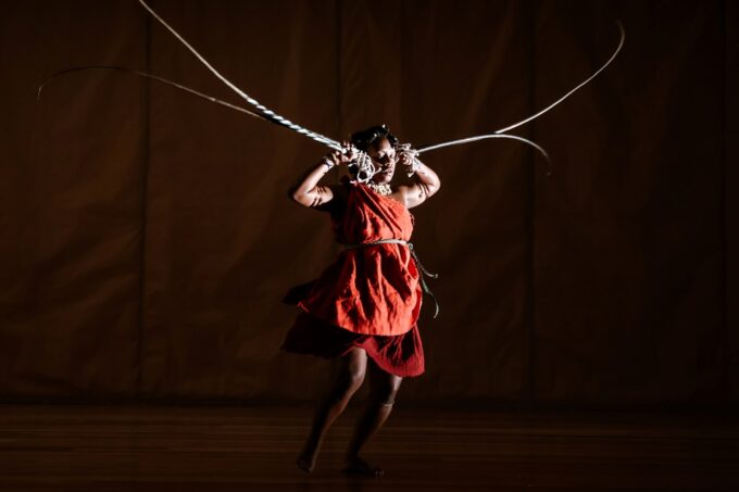 Photo of dancer performing, dressed in a red smock and holding long antler-like props by their ears.