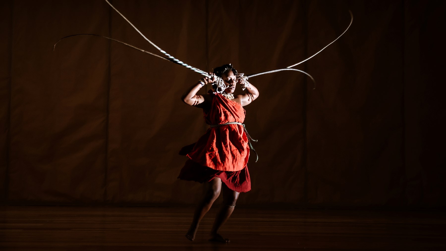 Photo of dancer performing, dressed in a red smock and holding long antler-like props by their ears.